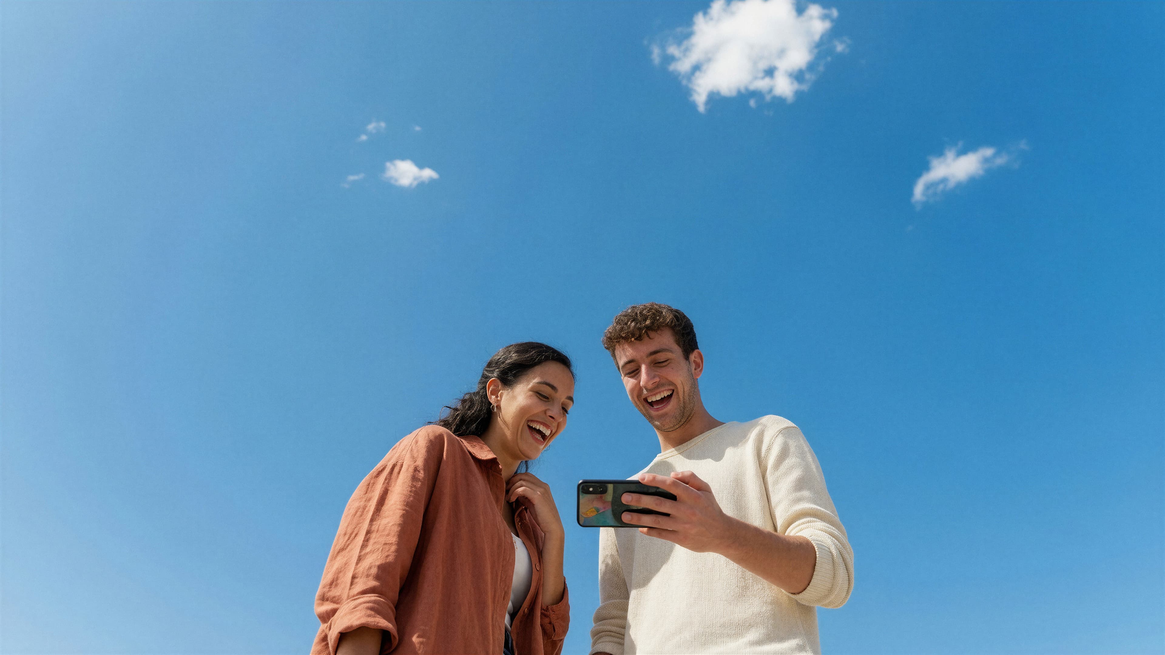 Dos personas sonriendo mientras miran un teléfono bajo un cielo azul.